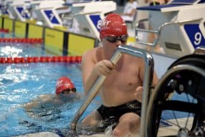 Mężczyzna wychodzi z wody. Za nim czeka kolejny. Przed sobą ma wózek inwalidzki.