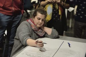 Kucająca kobieta, trzyma długopis nad kartką. Drugą ręką podpiera sobie kark. Za nią stoją ucięci na fotografii ludzie.