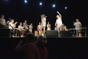 Na scenie jest grupa ludzi. Na środku dwie kobiety stoją i trzymają ręce w górze. Za nimi w półkolu siedzą osoby i niektórzy mają przed sobą bębny.