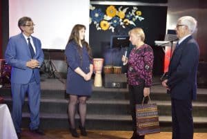 Ludzie stoją na scenie. Twarze mają zwrócone ku sobie. Jedna z kobiet trzyma w dłoni torbę upominkową.