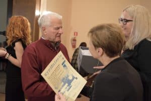 Mężczyzna stoi przed dwiema kobietami. Jedna z nich się uśmiecha a druga trzyma kartkę z wyróżnieniem. Obie stoją bokiem.