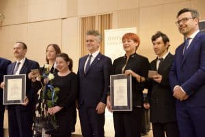 Kobiety i mężczyźni na scenie pozują do zdjęcia. Trzymają oni kwiaty oraz dyplomy.