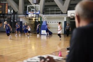 Kobieyu grają na boisku w koszykówkę. W tle jest widownia. Mędzyny z widowni ma twarz skierowana w stronę boiska.