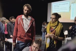 Dwójka młodych ludzi , kobieta i mężczyzna przyglądają się pracy innej kobiety, która jest ucięta na dole fotografii. Kobieta patrzy na punkt, który nie został umieszczony na fotografii.