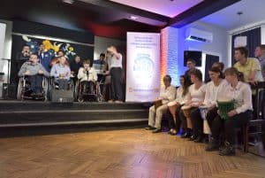 Ludzie na wózkach inwalidzkich są na scenie. Po lewej stronie ludzie siedzą na krzesłach. Między nogami maja bębenki.