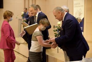 Mężczyzna gratuluje dziewczynce i wręcza jej pudełeczko. Obok stoi mężczyzna z biała róża. Po lewej stronie starszy mężczyzna, który trzyma dyplom wyciąga rękę do kobiety, która idzie.