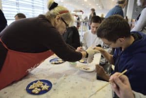 Kobieta w fartuchu pochyla sie w stronę chłopca siedzącego przy stole. Trzyma go za dłoń.