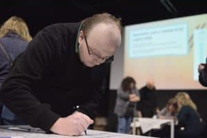 Pochylony mężczyzna w średnim wieku, trzymając w prawej dłoni długopis uzupełnia dokumenty leżące na blacie. W tle wyświetlana jest prezentacja.