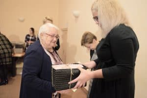 Starsza kobieta stoi a na przeciwko jest kobieta z paczką wyciągniętą w jej kierunku. Za nimi stoi kobieta. Wszyscy stoją bokiem do obiektywu.