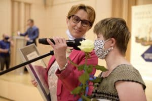 Dwie kobiety stoją przy mikrofonie. Kobieta po lewej stronie trzyma mikrofon a druga ma twarz blisko niego. Kobieta po prawej stronie trzyma róże i ma maskę na twarzy.