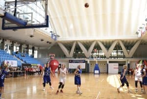 Osoby na boisku do koszykówki. Wysoko w górze jest piłka, a osoby patrzą w górę na piłkę.