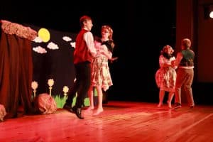 Na scenie jest grupa osób. Są to dwie pary, które lekko unoszą nad podłogą. Za nimi scenografia przedstawiająca łąkę.