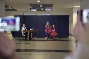 Ludzie stoją na scenie. Dwie kobiety w czerwonych sukienkach oraz mężczyzna siedzący za stołem.