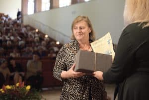 Kobieta stoi z paczką na scenie oraz dyplomem. Przed nią stoi kobieta tyłem i trzyma paczkę w geście dawania. Za kobietą jest widownia.