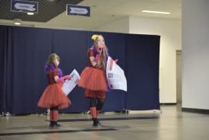 Dwie kobiety ze sceny w czerwonych sukienkach. Jedna z nich trzyma w dłoni mikrofon.