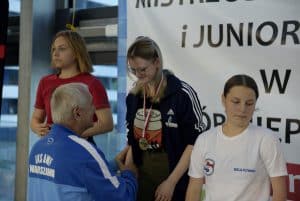 Trzy dziewczyny stoją. Starszy mężczyzna patrzy na dziewczynę po środku i wyciąga rękę w geście gratulacji.