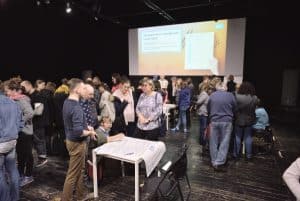 Wyświetlana jest prezentacja. Duża grupa ludzi swobodnie spaceruje między stołami, oddając się dyskusjom. Są wśród nich widoczne osoby niepełnosprawne m.in. na wózkach inwalidzkich.