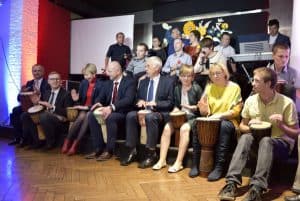 Ludzie na wózkach inwalidzkich są na scenie. Niektórzy trzymają grzechotki inni bębenki. Pod sceną siedza ludzie z bębenkami między nogami.