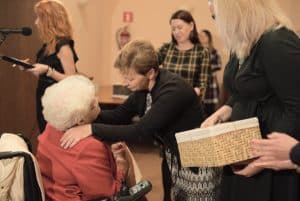 Kobieta siedzi na wózku inwalidzkim. Kobieta nachyla się do tej kobiety i ma ręce na jej barkach. Kobieta po prawej stronie trzyma koszyczek, a po lewej stoi kobieta przed mikrofonem.