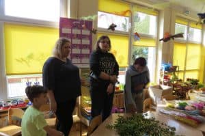 Trzy kobiety stoją przed stołem na którym jest bukszpan. Po lewej stronie siedzi chłopczyk na krześle. Z sufitu zwisają origami.