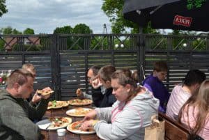 Przy dużym drewnianym stole jedna kobieta i czterech mężczyzn konsumują pizzę na tarasie.