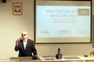 Starszy mężczyzna w okularach trzyma mikrofon i przemawia. Stoi obok wyświetlanej prezentacji.