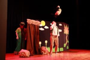 Na scenie są dwie osoby. Mężczyzna stoi lekko odchylony na nogach do tyłu. Dziewczyna stoi schowana za scenografia.