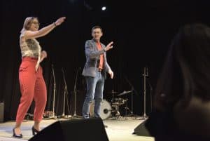 Kobieta i mężczyzna stoją na scenie. Kobieta ma jedną rękę wyciągniętą przed siebie. Mężczyzna stoi obok, patrzy na kobietę i także ma wyciągnięta rękę przed siebie.