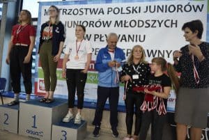 Trzy dziewczyny stoją na podium z medalami. Obok stoją kobiety, mężczyzna i dziewczynka. Dziewczynka trzyma futerał z medalami.