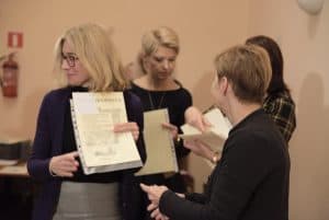 Kobiety stoją z kartkami w dłoniach. Żadna nie patrzy w obiektyw. Kobieta po prawej stronie stoi bokiem.