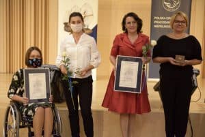 Cztery kobiety pozują do zdjęcia. Jedna z nich jest na wózku inwalidzkim. Dwie kobiety trzymają dyplomy, a pozostałe różę lub pudełeczko.