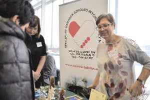 Kobieta stoi przy stoisku reklamowym. Uśmiecha się. Za nią pionowy baner reklamowy.