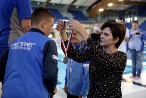 Kobieta trzyma medal. Chłopak nachyla się ku niemu. Obok stoją inni ludzie.