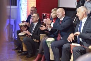 Ludzie siedzą pod sceną z bębnami między nogami. Uśmiechają się.