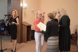 Na scenie stoją kobiety. Po prawej stronie kobieta trzyma wyróżnienie, a przed nią stoi kobieta. Obok nich stoją kobiety. Po lewej stronie kobieta stoi przed mikrofonem a głowę ma zwróconą na grupę kobiet.