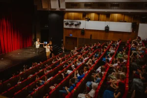 Osoby na widowni oglądają co dzieje się na scenie. Na scenie stoi mężczyzna.