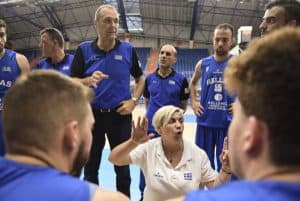 Grupa sportowców stoi w kole. Pośrodku kobieta siedząca w kucki ma podniesione ręce, usta ułożone w dzióbek.