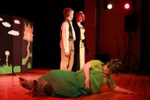 Na scenie są trzy osoby. Jedna osoba leży na scenie, a dwie stoją nad nią. Za nimi scenografia.