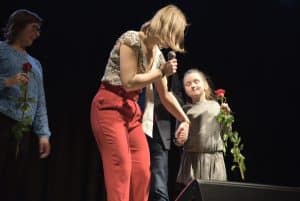 Kobieta stoi z mikrofonem a głowę odwraca w stronę dziewczynki, która trzyma różę. Za nimi stoi kobieta z różą.