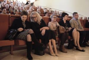 Duża grupa osób siedzi na widowni. Na przodzie siedzą cztery kobiety i mężczyzna. Kobieta nachyla się do drugiej.