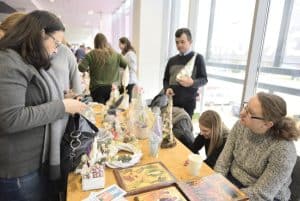 Ludzie siedza przy stole z rękodziełami. Niektórzy stoją i pochylają sie w ich kierunku.