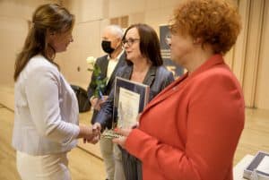 Kobieta po prawej stronie stoi i trzyma statuetkę. Obok dwie kobiety patrzą na siebie i wykonują dłońmi gest gratulacji. Za nimi starszy mężczyzna trzyma różę.