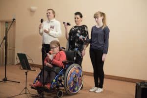 Trzy kobiety i mężczyzna są na scenie. Z przodu jest kobieta na wózku inwalidzkim i ma mikrofon blisko twarzy. Za nią stoją trzy osoby a w rękach mają grzechotki.