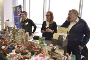 Ludzie stoją przy stoisku z rękodziełami. Ich twarze są zwrócone na wprost.