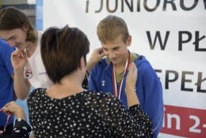 Kobieta stoi przed chłopcem. ma on na sobie medal. Kobieta trzyma przy nim ręce jakby poprawiała medal.