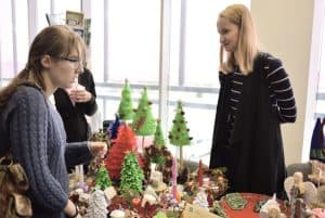 Kobieta stoi przy stoisku z rękodziełami, wśród których są choinki i anioły. Inna kobieta pochyla się w kierunku tego stolika.