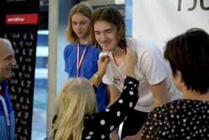 Chłopak nachyla się do kobiety. Ona trzyma ręce na wysokości jego szyi jakby poprawiała jeszcze medal. Po lewej stronie stoi starszy mężczyzna.