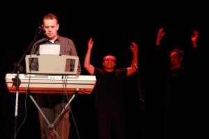 Na scenie stoi mężczyzna przed keyboardem. Za nim po prawej stronie stoją mężczyźni, którzy mają podniesione ręce.