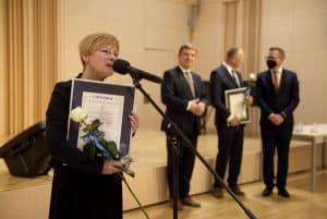 Kobieta z dyplomem i różą przemawia do mikrofonu. Z boku po prawej stronie stoją trzej mężczyźni. Jeden nich także ma dyplom i różę.