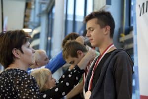 Kobieta stoi przed chłopakiem i patrzy na niego. On ma zamknięte oczy i medal na sobie. Za nimi kobieta nakłada medal innemu chłopcu.
